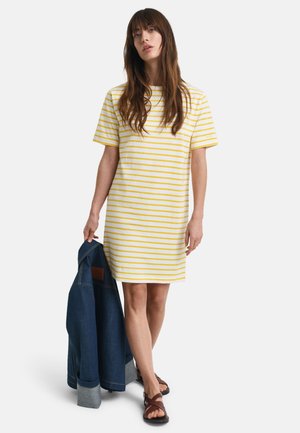 Striped kjole med korte ermer i hvit og gul, med en løs passform. En denimjakke holdes i den ene hånden, og brune sandaler er på føttene.
