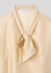 Blusa beige chiaro con una consistenza morbida e simile alla seta. Presenta un nodo al collo e una leggera lucentezza, progettata per una silhouette aderente. Etichetta del marchio visibile.
