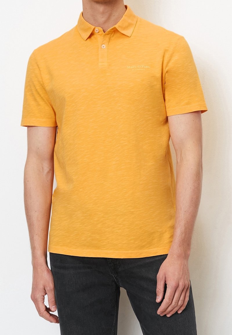 Homme portant un polo à manches courtes orange vif et un jean foncé, debout devant un fond clair uni.