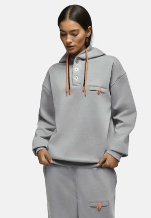Femme portant un sweat à capuche gris et un pantalon assorti avec des cordons orange et des détails de boutons, debout les mains près des poches.