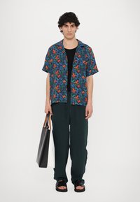 Short-sleeve, blue floral shirt with red and green patterns, paired with loose, dark green pants and a black and white tote bag. Black sandals.