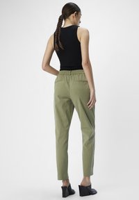 Pantalon tapered vert clair avec une taille élastique, muni de poches arrière. Porté avec un haut sans manches noir et des mules à talons noires.