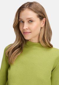 Fiatal nő világosbarna hajjal, lime zöld pulcsit visel, enyhén oldalra nézve, semleges arckifejezéssel.