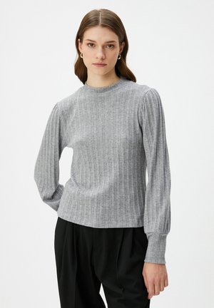Eine Frau trägt einen grauen, gerippten Pullover mit puffartigen langen Ärmeln und schwarze Hosen, und steht vor einem schlichten weißen Hintergrund.