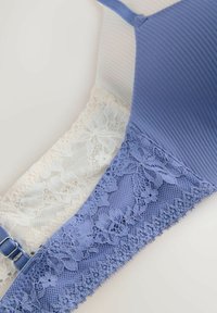 Sutiene albastre și albe cu detalii florale din dantelă și material cu dungi, așezate pe o suprafață deschisă la culoare, prezentând bretelele și încuietorile reglabile.