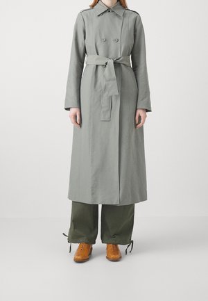 Personne portant un trench-coat gris ceinturé, un pantalon large vert avec des liens aux chevilles et des chaussures marron à lacets, debout devant un fond uni.