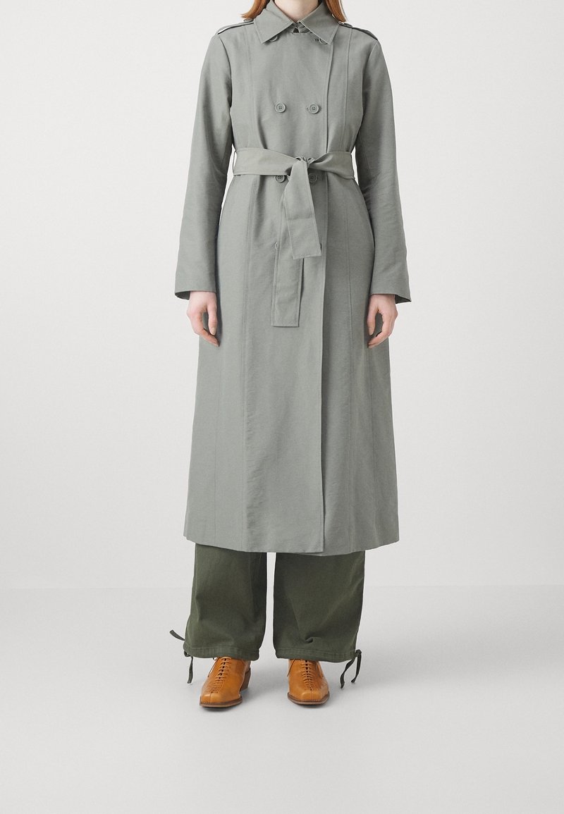 Personne portant un trench-coat gris ceinturé, un pantalon large vert avec des liens aux chevilles et des chaussures marron à lacets, debout devant un fond uni.