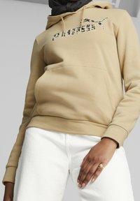 Sudadera con capucha beige con un bolsillo canguro en el frente, que presenta un logo de Puma en blanco y negro. Combinada con jeans blancos y un anillo en la mano.