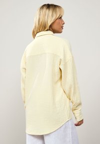 Yellow textured shirt with a collared neckline and long sleeves. The fabric has a subtle raised pattern and a curved hemline.