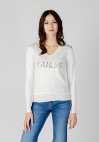 Maglione bianco con scollo a V e maniche lunghe. Presenta la scritta "GUESS" in lettere decorate sulla parte anteriore. Indossato con jeans blu.