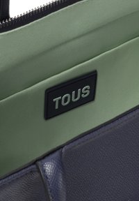 Interior de un bolso de cuero texturizado en color azul marino con forro de tela verde claro; presenta una etiqueta negra con "TOUS" en letras plateadas.
