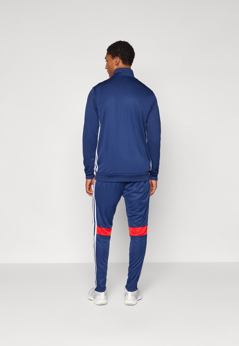 Chándal azul marino compuesto por una chaqueta con cremallera y pantalones, con rayas blancas en las mangas y acentos rojos en las piernas.