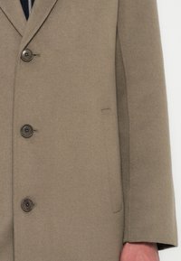 Beige gyapjú kabát, kihajtott gallérral, négy gombbal záródik, és oldalsó zsebe van, sima, textúrált felülettel.