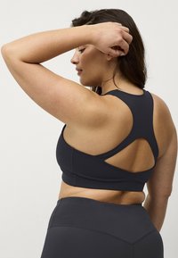 Sort sports-bh med racerback-design og udskæringsdetalje. Lavet af strækbart materiale, der giver en tætsiddende og støttende silhuet.