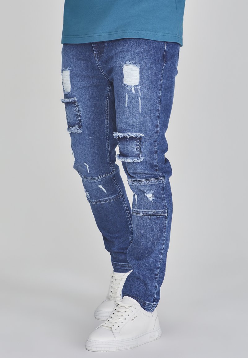 Ripped blue denim jeans featuring distressed patches, frayed edges, and a tapered fit. Paired with white sneakers.