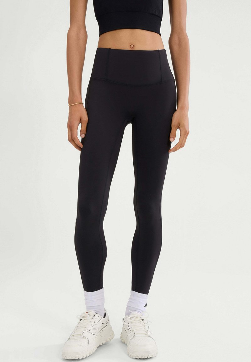 Hoge zwart leggings van rekbare stof, met een gladde textuur en naaddetails, gecombineerd met witte sneakers en sokken.