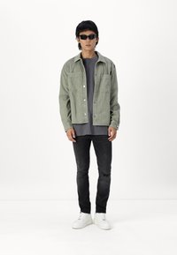 Chaqueta de camisa verde claro con dos bolsillos, superpuesta a una camisa gris oscuro. Jeans negros con un detalle deshilachado, combinados con zapatillas blancas.