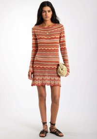 Robe en tricot dans des tons chauds de rouge, orange et crème ; présente un motif ondulé, des manches longues et un ourlet festonné ; associée à une pochette texturée.