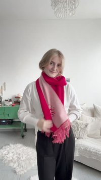 Νεαρή γυναίκα που φτιάχνει ένα κόκκινο και ροζ φουλάρι με κρόσσια, στέκεται σε ένα φωτεινό σαλόνι με πράσινη ντουλάπα και λευκό καναπέ.