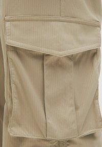 Beżowe spodnie cargo mają pionowe paski, dużą boczną kieszeń z klapą oraz wzmocnione szwy. Tkanina jest gładka z delikatnym połyskiem.