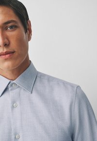 Light blue textured shirt with a button-down collar, featuring a subtle pattern and light buttons, shown in a close-up on a model's upper body.