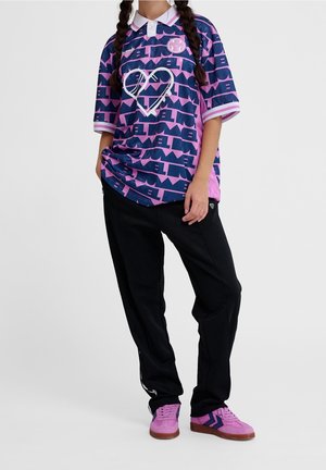 Osoba ubrana w fioletowo-granatową koszulę z krótkim rękawem w wzór z sercem, czarne spodnie i fioletowe trampki, stojąca z rękami w kieszeniach.