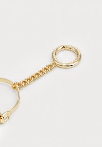 Porte-clés en or avec un design à maillons, un anneau circulaire et un logo de marque gravé. Texture lisse avec une finition métallique brillante.