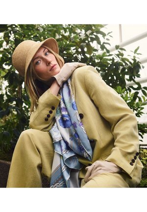 Woman in beige suit, floral scarf, beige gloves, and straw hat poses thoughtfully amid green plants.