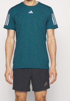 Mężczyzna ubrany w turkusową koszulkę Adidas z krótkim rękawem z białymi paskami oraz czarne spodenki Adidas stojący na jednolitym tle.
