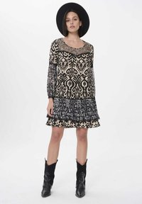 Schwarz-creamfarbenes gemustertes Kleid mit durchsichtigen langen Ärmeln, gerüschtem Saum und tailliertem Oberteil; kombiniert mit schwarzen Cowboystiefeln und einem breitkrempigen Hut.