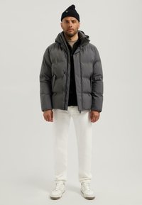 Grijze gewatteerde jas met een capuchon, zwart knoopshirt eronder, witte broek en witte sneakers. De stof lijkt glad en warm.