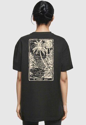 Schwarzes Baumwoll-T-Shirt mit einem großen beigen Grafikmotiv, das Palmen und einen Sonnenuntergang über Wasser zeigt, eingerahmt von einem rechteckigen Rand.