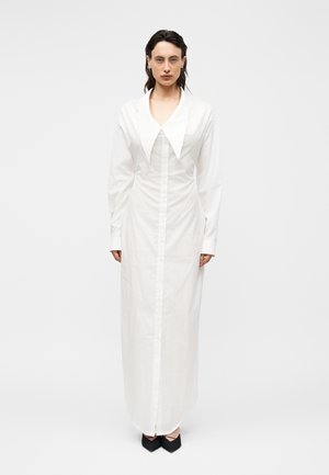 Frau trägt ein langärmeliges, bodenlanges weißes Kleid mit Knöpfen, einem übergroßen Kragen und schwarzen spitzen Schuhen, steht vor einem einfarbigen Hintergrund.