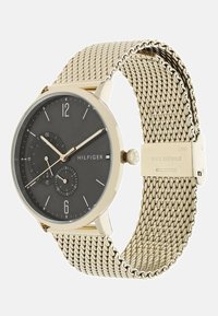 Montre noire ronde avec un bracelet en maille acier inoxydable doré, affichant des marqueurs et aiguilles argentés. Caractéristiques de doubles sous-cadrans pour l'heure au format 24 heures.