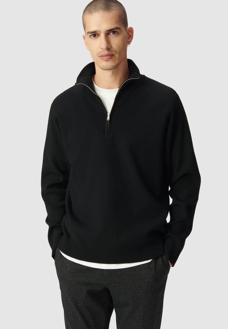 Czarny dzianinowy sweter z zamkiem błyskawicznym przy kołnierzu, z prążkowaną teksturą na ramionach i mankietach, noszony na białej koszuli.