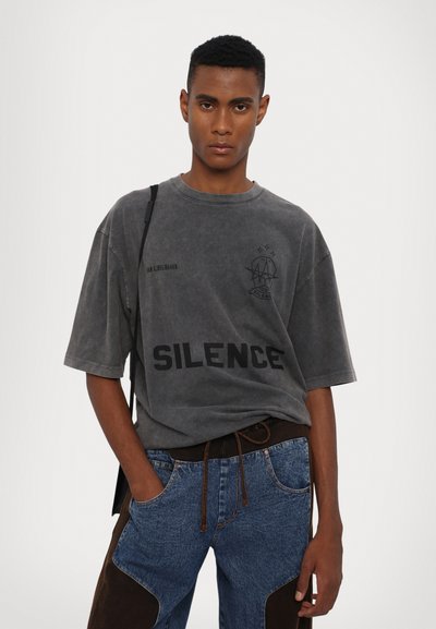 T-shirt oversize gris avec "SILENCE" imprimé en noir. Présente un col rond et des manches courtes, associé à un jean en denim bleu et des accents marron.