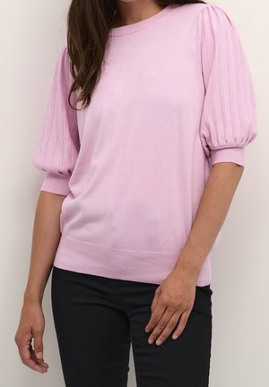 Woman wearing a light pink knit top with puffed ribbed sleeves and black pants, standing against a plain light background.