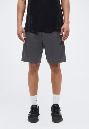 Mężczyzna w ciemnoszarych szortach Adidas, czarnej koszulce, białych skarpetach Adidas i czarnych butach sportowych Adidas, stojący na jednolitym tle.