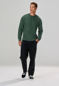 Long-sleeve green shirt in cotton blend, relaxed fit. Paired with black baggy jeans and black-and-white sneakers. Plain design, no patterns.