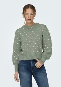 Lichtgroene gebreide trui met een getextureerd bobblepatroon en ribgebreide manchetten, gecombineerd met blauwe jeans. Eenvoudige ronde halslijn en cropped ontwerp.