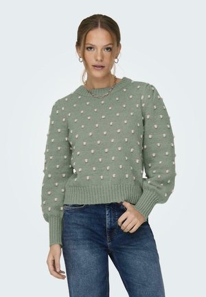 Hellgrüner Strickpullover mit strukturiertem Bobbelmuster und gerippten Bündchen, kombiniert mit blauen Jeans. Schlichte runde Ausschnittform und cropped Design.