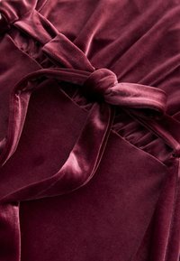 Mīksta bordo samta auduma ar sasaistītu loku detaļu. Tekstūra ir maiga un gluda, ar elegantiem drapējumiem un sakopotām daļām.