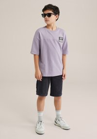 Levanduľové tričko s krátkym rukávom a nášivkou na hrudi, skombinované s tmavými šortkami. Model má na sebe čierne slnečné okuliare a šedé pruhované ponožky s bielymi teniskami.