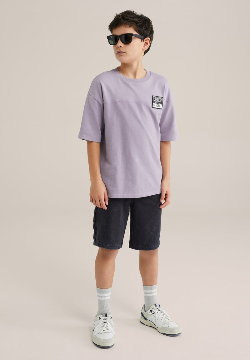 Levanduľové tričko s krátkym rukávom a nášivkou na hrudi, skombinované s tmavými šortkami. Model má na sebe čierne slnečné okuliare a šedé pruhované ponožky s bielymi teniskami.