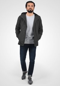 Veste légère noire à capuche, sweat-shirt gris en dessous, jean foncé et baskets grises. Comprend une fermeture éclair et deux poches latérales.