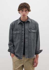 Camicia di jeans grigio scuro con maniche lunghe, chiusura con bottoni, due tasche sul petto e vestibilità comoda, indossata sopra un top nero e pantaloni color khaki.