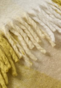 Uldent tæppe med en gradient fra bleg gul til hvid, med lange, frynsete frynser og en blød, tekstureret overflade.