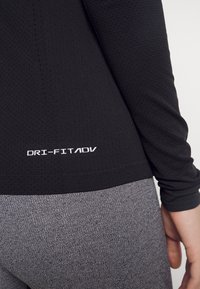 Maglietta sportiva nera a maniche lunghe con tessuto testurizzato, dotata di un logo bianco "DRI-FIT ADV" vicino all'orlo. Abbinata a leggings grigi.
