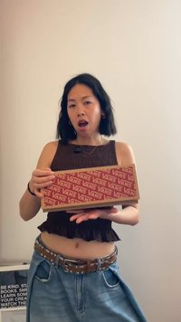 Νεαρή γυναίκα με κοντά μαύρα μαλλιά κρατά ένα κόκκινο κουτί παπουτσιών Vans, στέκεται μπροστά σε έναν απλό φωτεινό τοίχο φορώντας καθημερινά ρούχα.