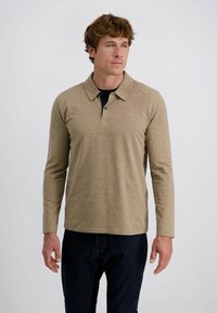 Mann mit lockigem Haar, der ein beiges Langarm-Polohemd und dunkle Jeans trägt und vor einem einfarbigen, hellen Hintergrund steht.